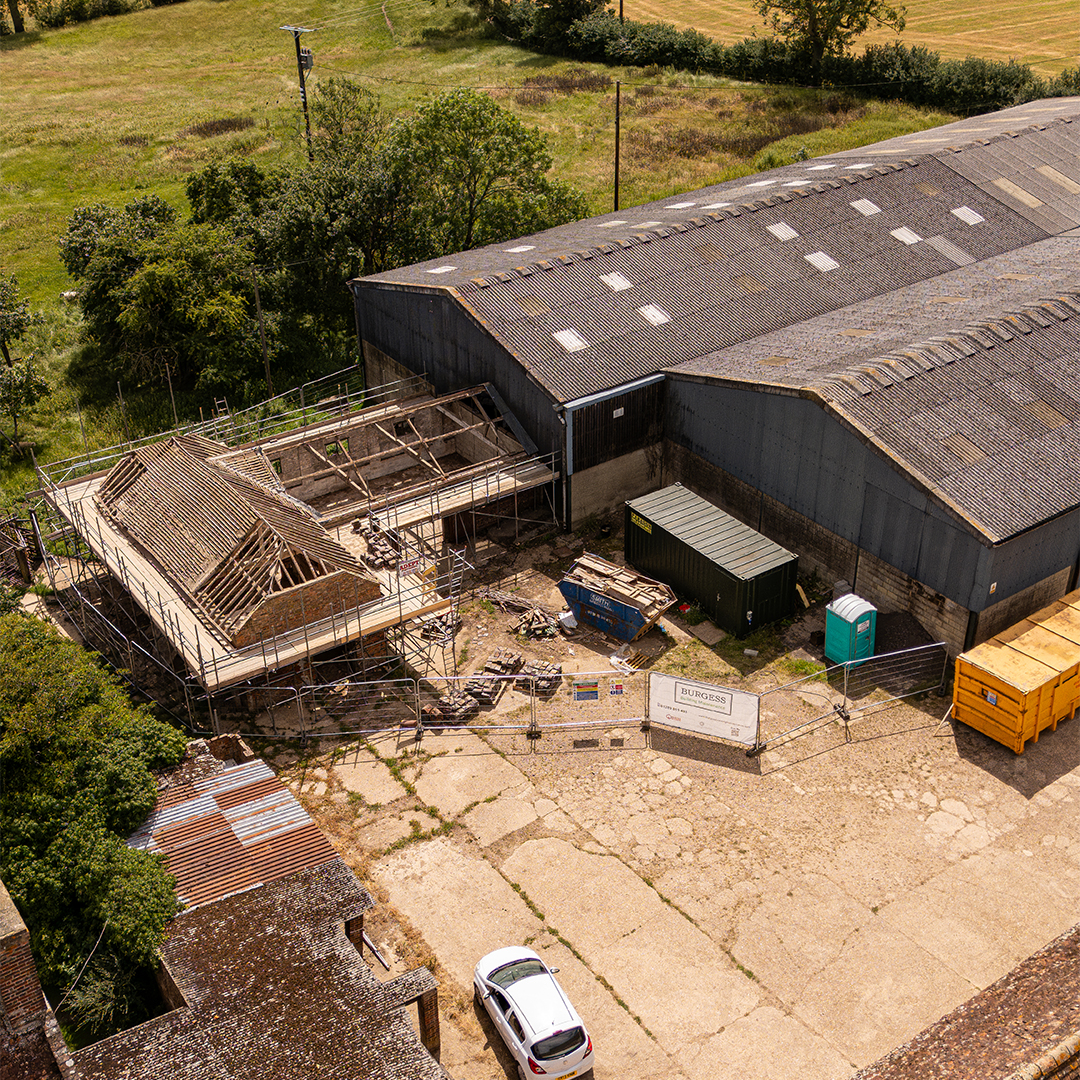 Aerial drone view of a rural building renovation, showing scaffolding, exposed timber roof structures and an active construction site beside agricultural barns.