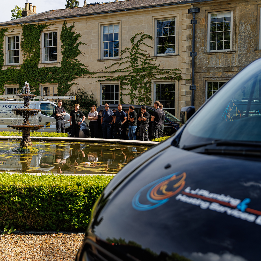 L J Plumbing & Heating Services Ltd team gathered beside branded work vans outside a large stone property with a landscaped fountain.