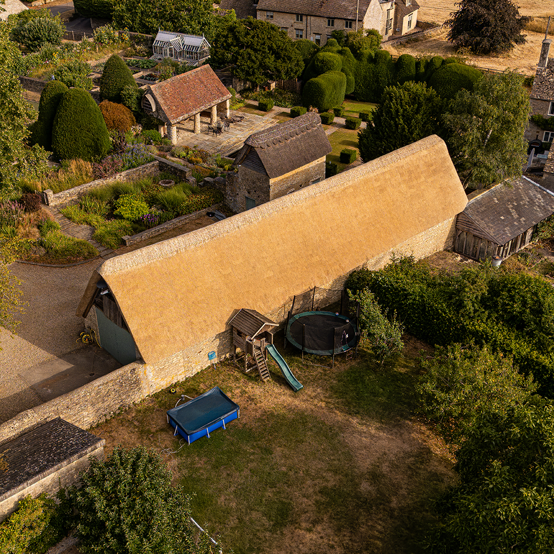 Aerial drone view of a newly thatched barn surrounded by traditional stone buildings, landscaped gardens and mature trees.